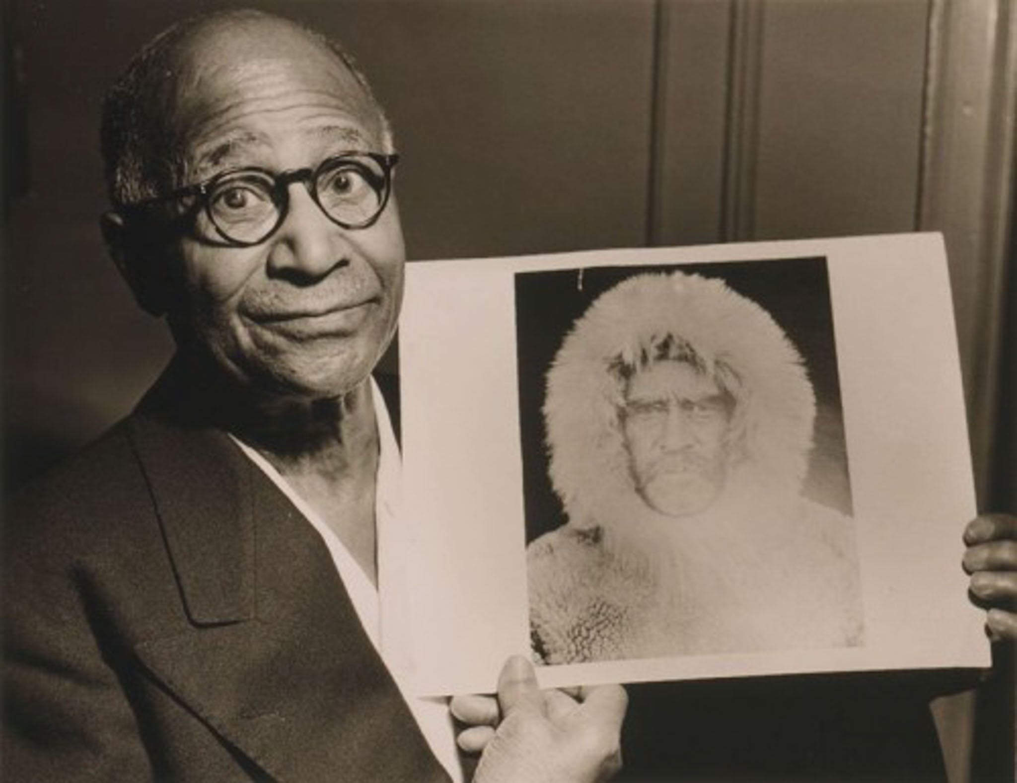 A man holds a photograph of a man wearing a fur coat with hood.