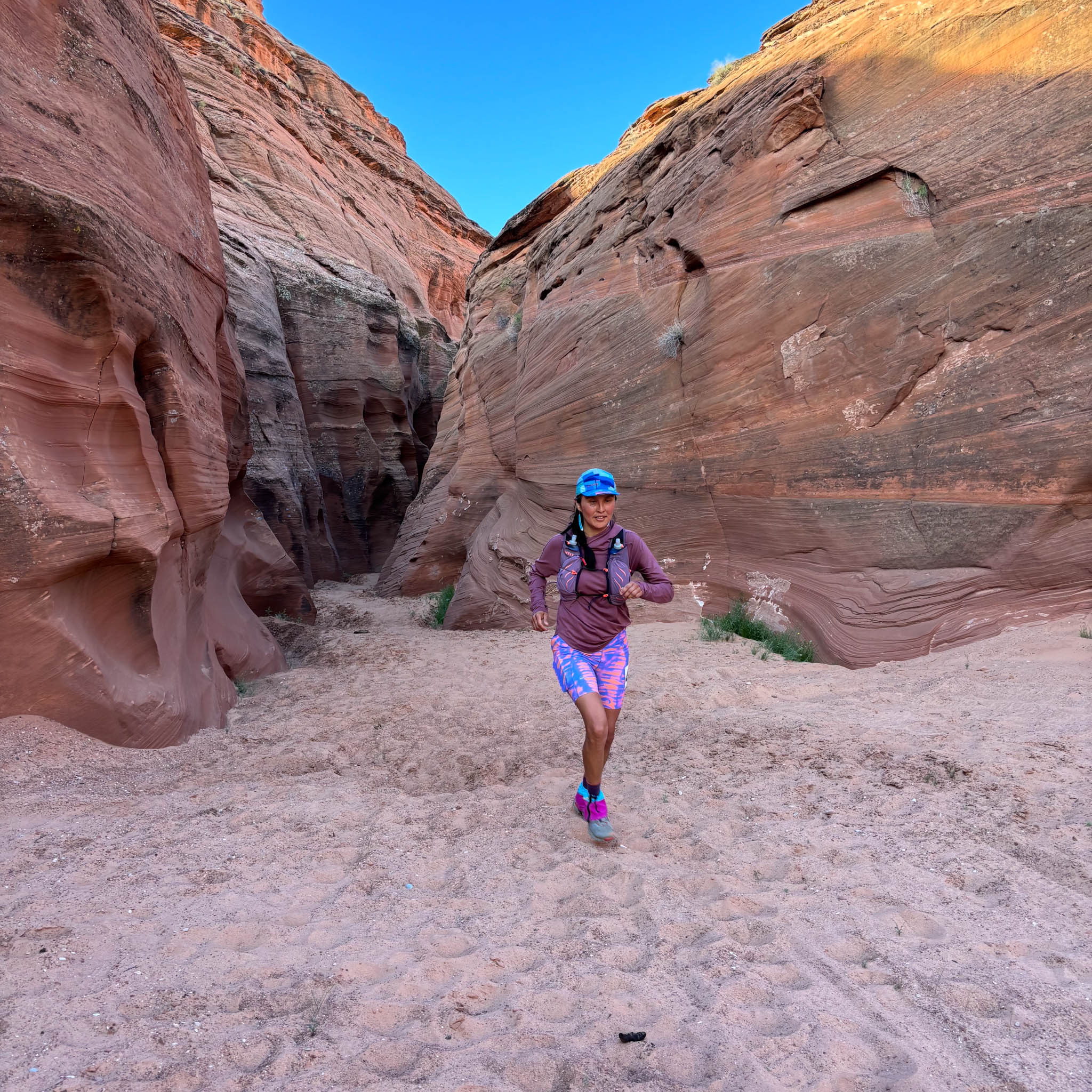 A person runs on a sandy trail from a narrow canyon entrance.
