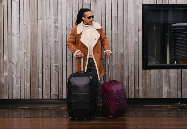 A person in a brown coat holding two wheeled suitcases, one black and one maroon, while standing in front of a wooden wall.