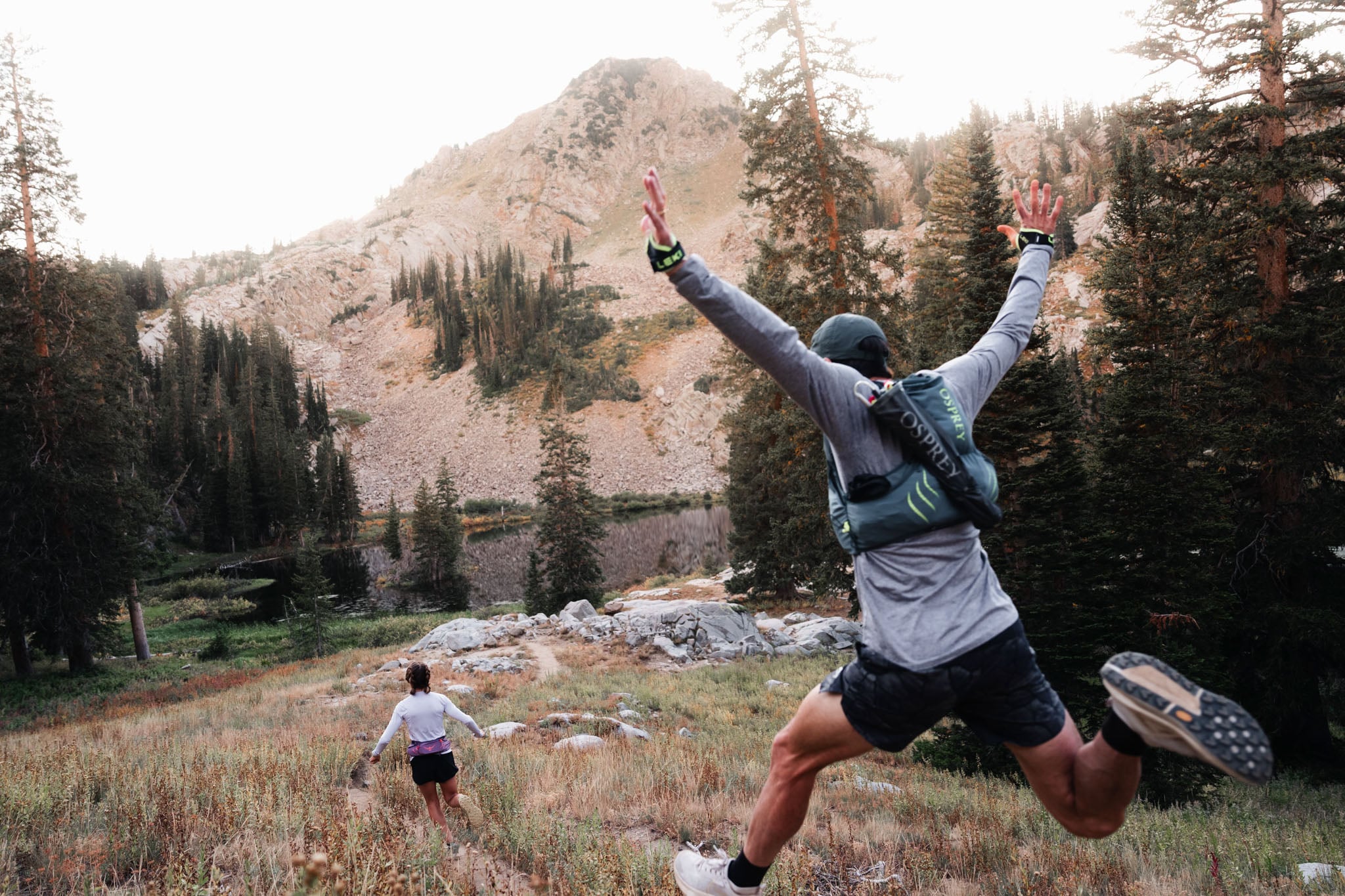 Two people run away from the camera in the mountains. The person closest to the camera leaps.