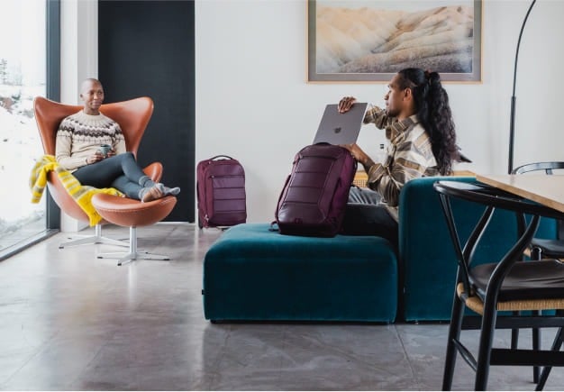 Two people relaxing indoors, one sitting in an orange chair and the other using a laptop on a teal sofa, with maroon travel backpacks placed beside them.