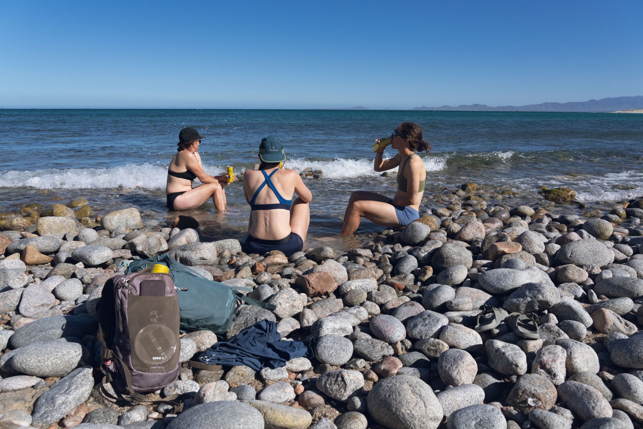 Three people sit in a pool by the sea shore drinking from cans.