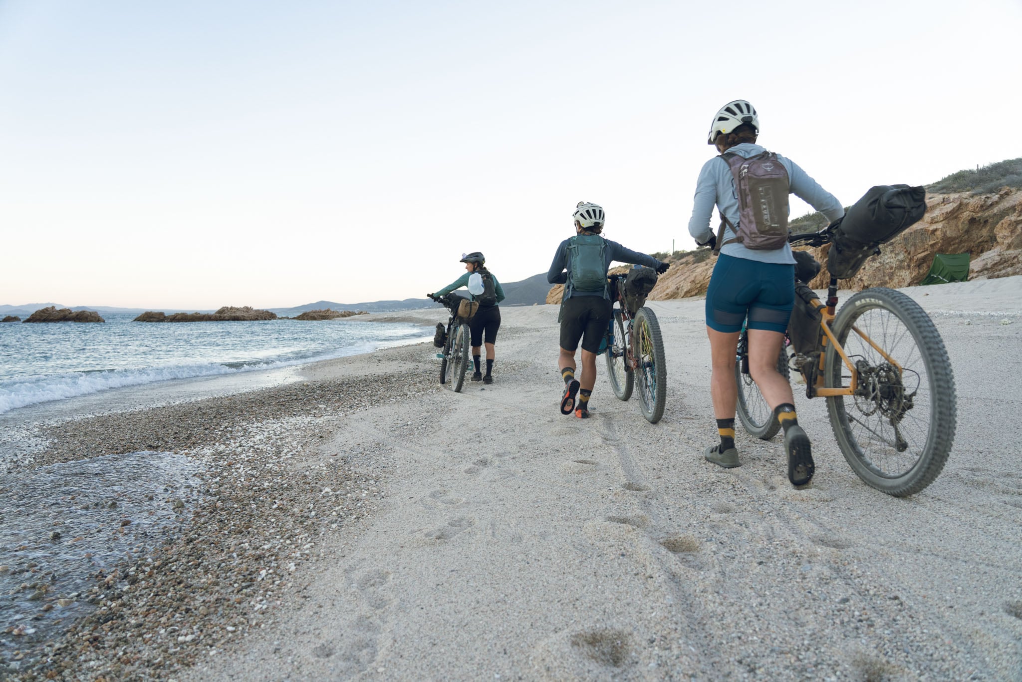 Three cyclists walk their bikes down the shore.