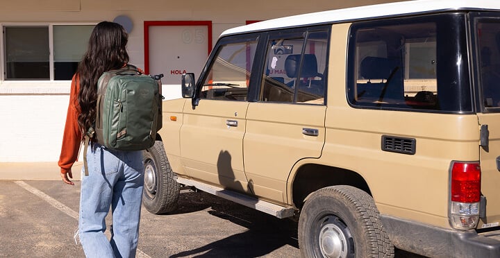Person wearing an Osprey Transporter 26+6 Expandable Travel Backpack walking next to a beige vehicle parked in front of a motel.