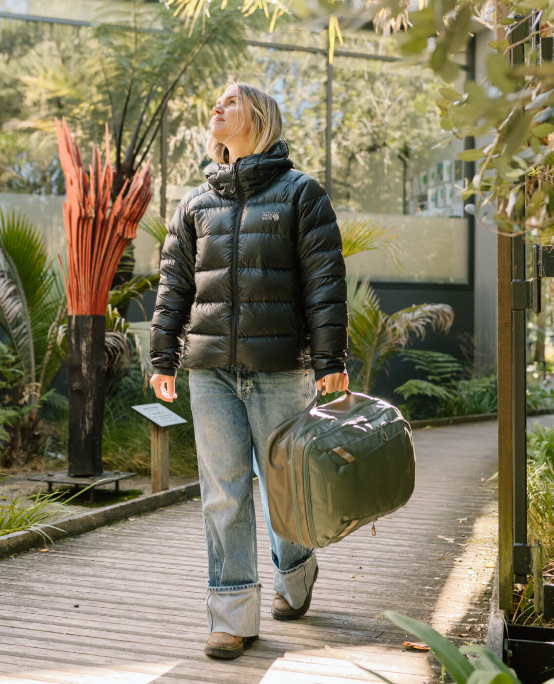 A woman walks down a garden path carrying a travel bag in her hand.