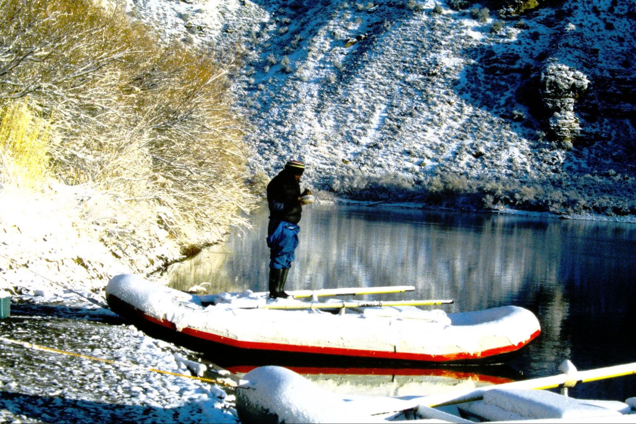 A man eats from a bowl on the edge of a river next to a raft.