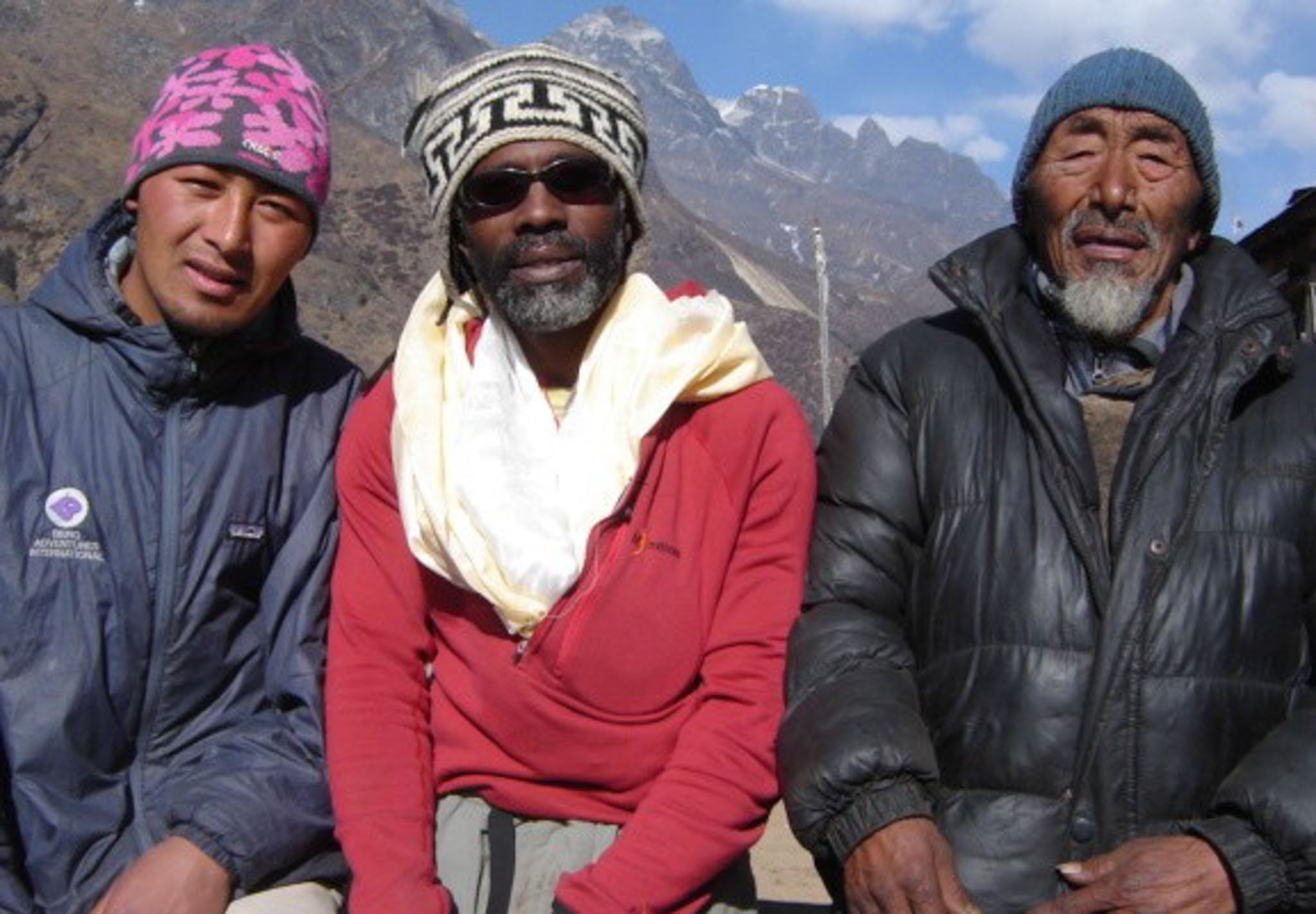 Three men in hats pose in front of the camera with mountains in the background.