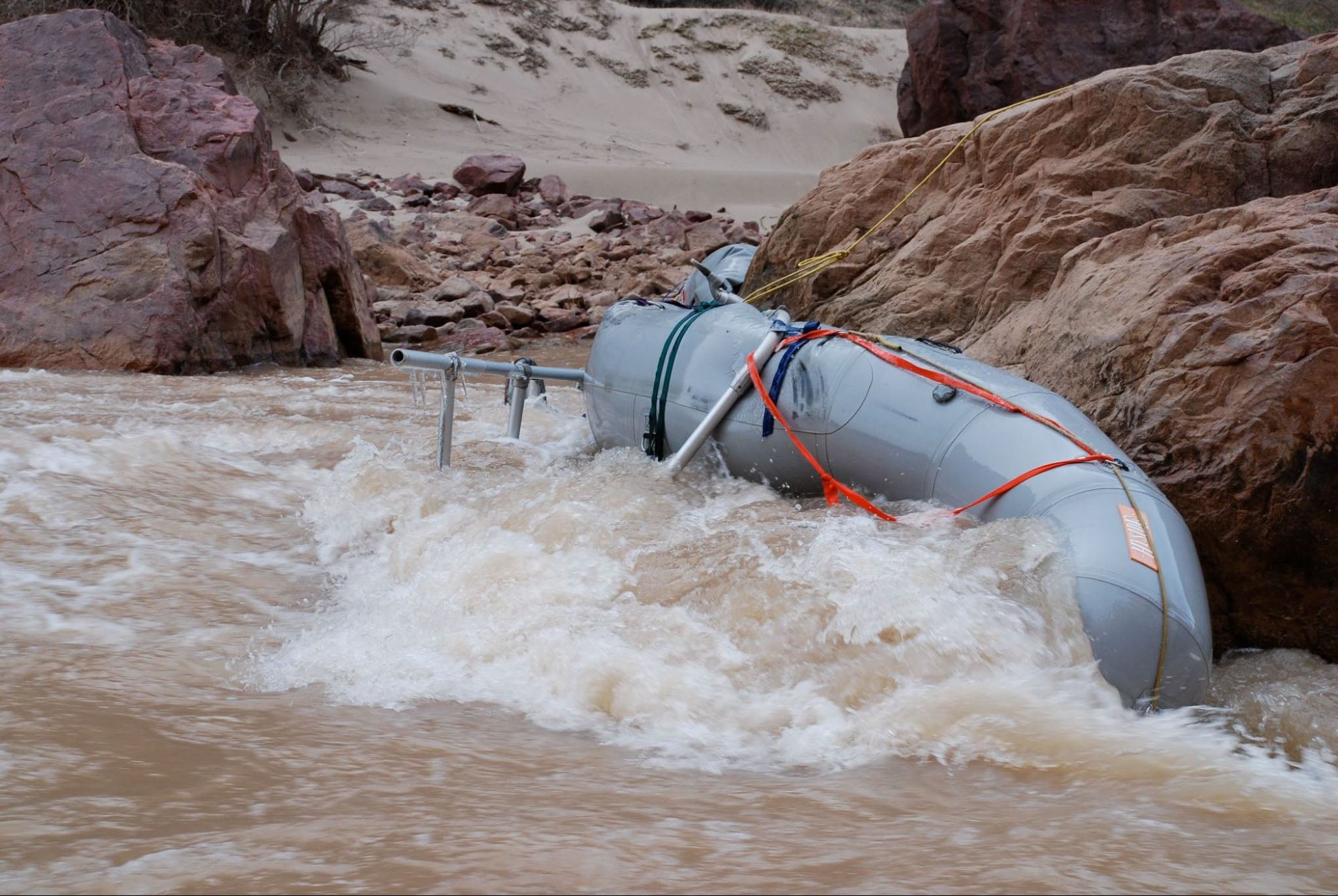 An inflatable raft is wrapped around a rock with waves of the river pushing it tight to the rock.