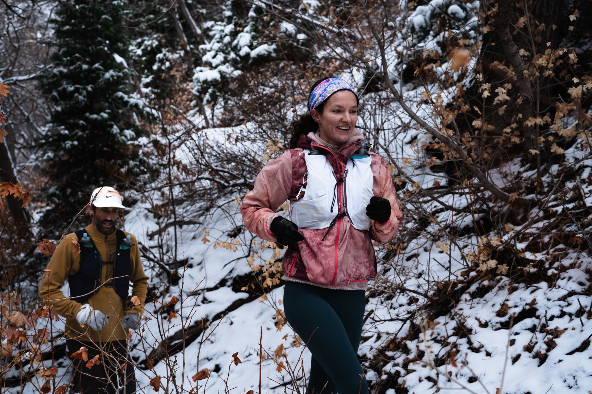 Two people run on a snowy forested trail.