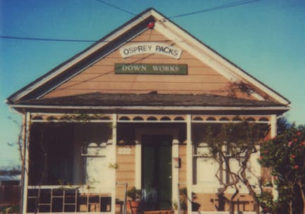 Faded, vintage photo of a peach-taupe Craftsman-style house with a front porch, converted into a storefront with two signs that say, &ldquo;Osprey Packs&rdquo; and &ldquo;Down Works.&rdquo;