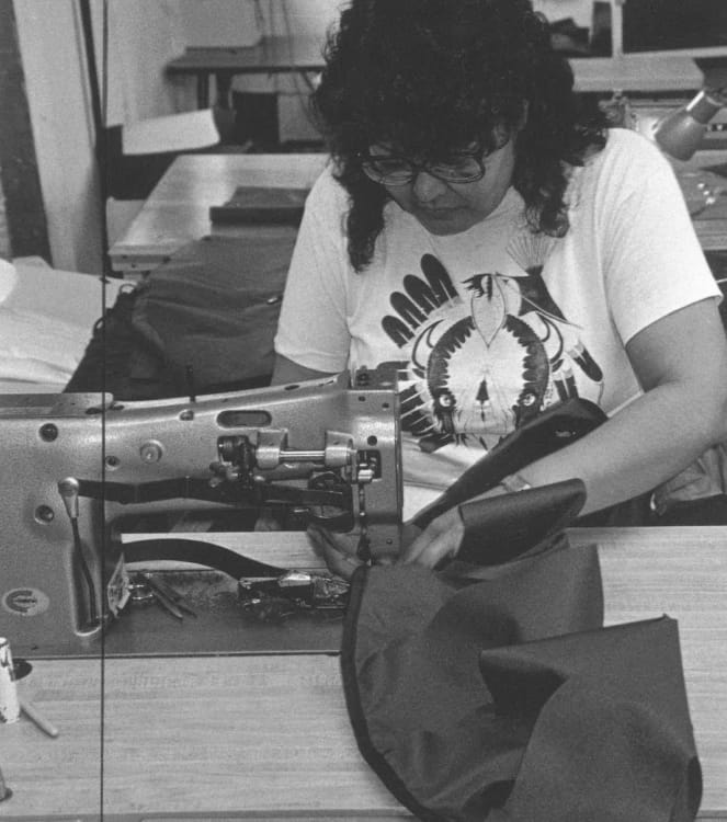 A black and white photo of a woman with curly black hair, glasses, and a white graphic t-shirt focused on sewing backpack components using a machine.