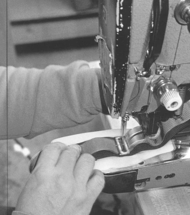 A close-up black and white photo of hands maneuvering a sewing needle around metal hardware on a backpack strap.