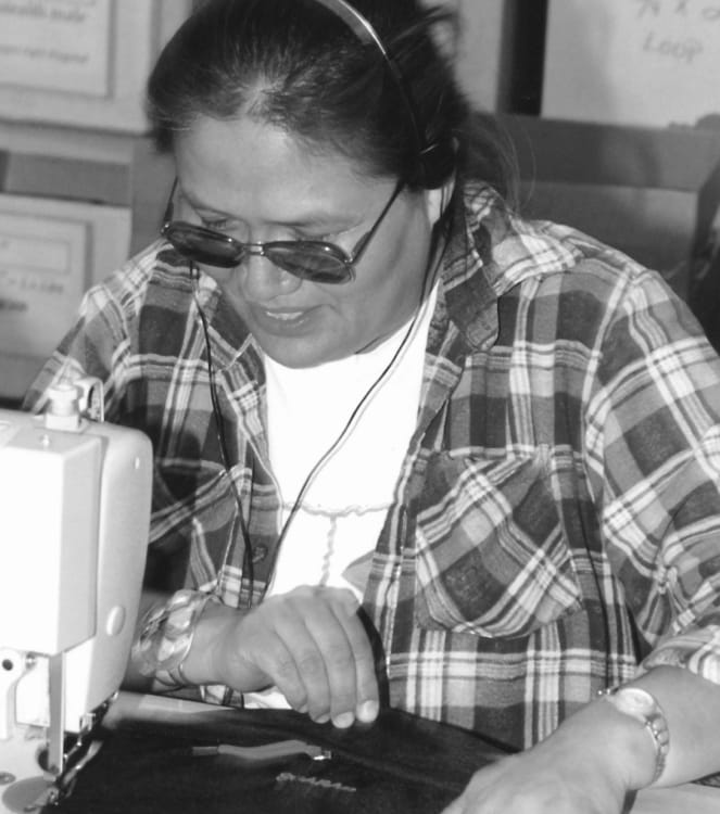 A black and white photo of a smiling woman who is sewing backpack components using a machine and wearing sunglasses and headphones.