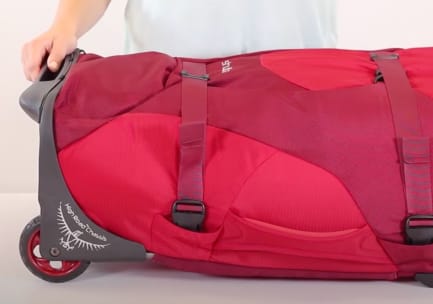 A red wheeled bag is laying on its back. Two StraightJacket compression straps with padded sidewalls are cinched tightly over the main body. The wheel frame bears an Osprey insignia and reads, &ldquo;High Road Chassis.&rdquo; A person in a white t-shirt grips the chassis.