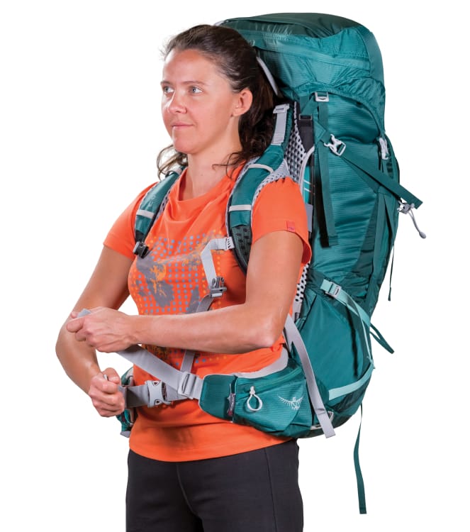 A woman in an orange graphic t-shirt wearing a teal backpacking pack over her shoulders is tightening the hipbelt&rsquo;s nylon
adjuster strap.