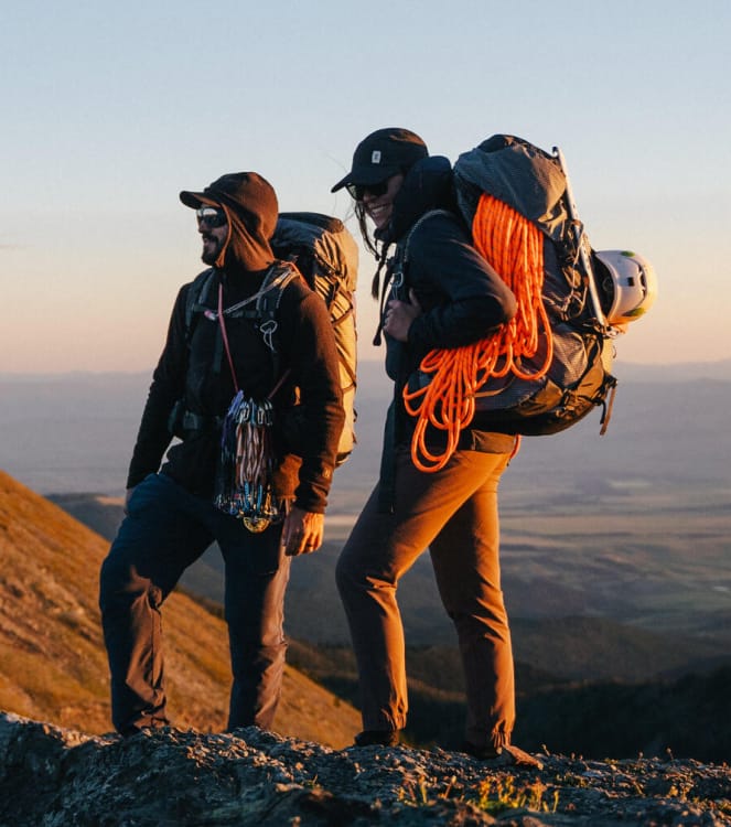 Two climbers are standing in morning light on a mountain ridge, wearing fully-loaded packs with a climbing rope, carabiners, helmets and an ice ax.