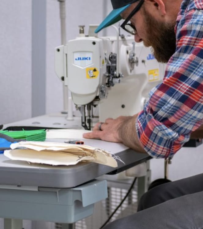 A bearded man wearing glasses, a baseball cap and a blue, white and red flannel shirt with rolled up sleeves constructs a cloth face mask on a sewing machine.