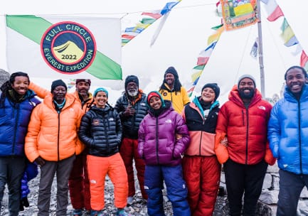 A team of seven climbers wearing
mountaineering pants and puffy coats under a white/green striped flag with a blue, red and yellow insignia that reads, &ldquo;Full Circle Everest Expedition 2022&rdquo; and Tibetan prayer flags blowing in the wind behind them.