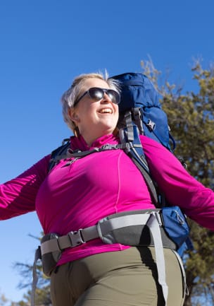 A photo of a silver-haired woman wearing a fuschia half-zip sweater, hiking pants, and a cobalt blue and gray Osprey backpack under a blue sky.