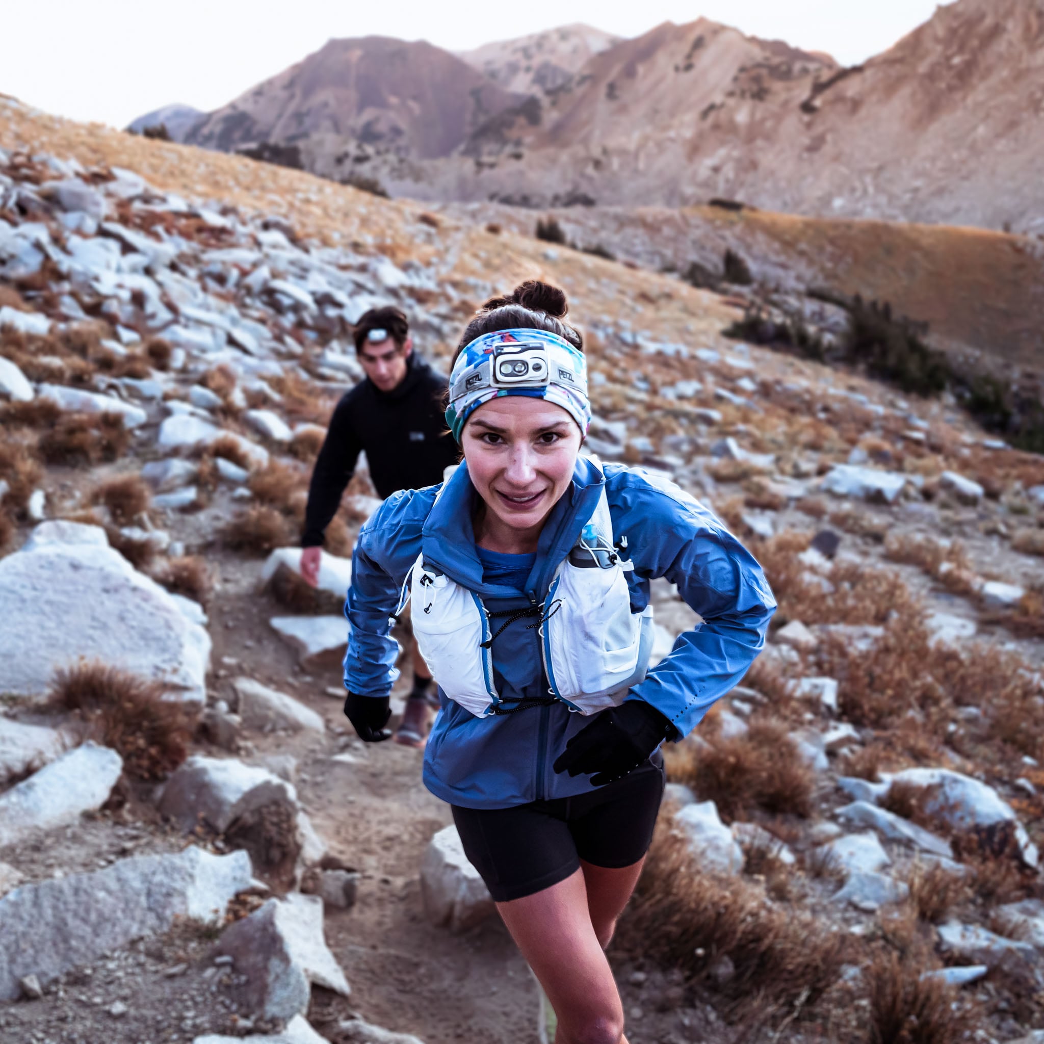 Two people run up a mountain trail toward the camera.