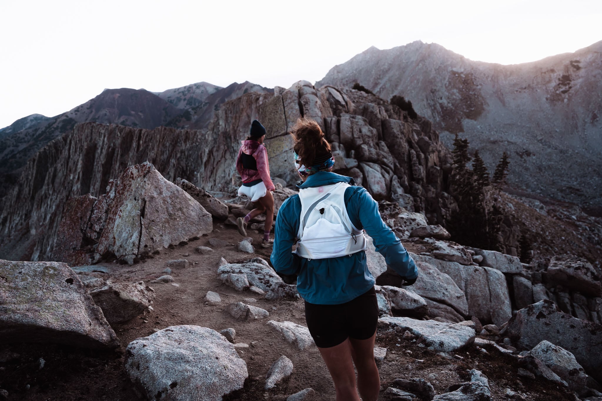 Two people run on a mountain trail away from the camera.
