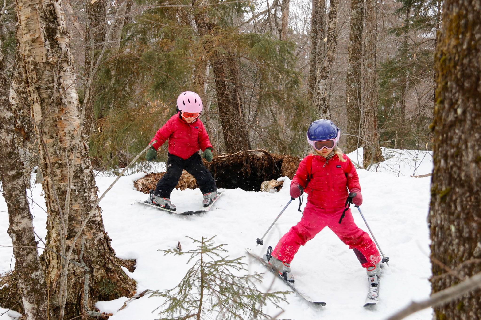 Two children in snow gear ski through the forest.